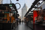 A modern indoor market or food hall with various stalls on either side. In the center, two people walk towards the entrance, where a unique, pyramid-shaped building can be seen through large glass windows.
