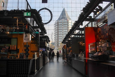 A modern indoor market or food hall with various stalls on either side. In the center, two people walk towards the entrance, where a unique, pyramid-shaped building can be seen through large glass windows.