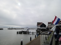A cloudy day at a waterfront with moored boats along a dock. Traditional buildings with dark facades and steep roofs line the shore. A flag with red, white, and blue stripes is visible. People appear to be walking or sitting on the dock.