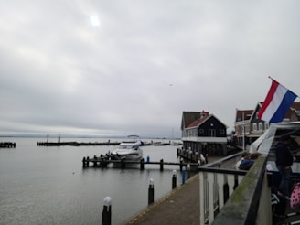 A cloudy day at a waterfront with moored boats along a dock. Traditional buildings with dark facades and steep roofs line the shore. A flag with red, white, and blue stripes is visible. People appear to be walking or sitting on the dock.