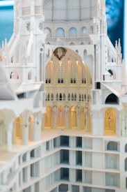An intricately designed architectural model showcasing detailed arches, columns, and vaulted ceilings. The model appears to represent a grand, multi-story structure with a white, ornate fa&ccedil;ade. The lighting highlights the detailed craftsmanship, revealing the interior's elegant and gothic-inspired design.