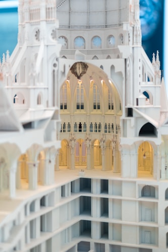 An intricately designed architectural model showcasing detailed arches, columns, and vaulted ceilings. The model appears to represent a grand, multi-story structure with a white, ornate fa&ccedil;ade. The lighting highlights the detailed craftsmanship, revealing the interior's elegant and gothic-inspired design.