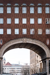 A detailed architectural view of a building facade with a geometric brick pattern. The upper part features rows of windows, some with arched tops, and the structure includes a large archway through which other urban elements, such as buildings and a church tower, can be seen.