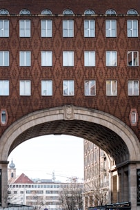 A detailed architectural view of a building facade with a geometric brick pattern. The upper part features rows of windows, some with arched tops, and the structure includes a large archway through which other urban elements, such as buildings and a church tower, can be seen.