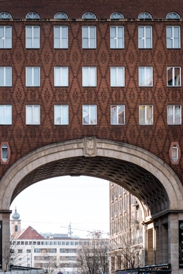 A detailed architectural view of a building facade with a geometric brick pattern. The upper part features rows of windows, some with arched tops, and the structure includes a large archway through which other urban elements, such as buildings and a church tower, can be seen.