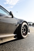 A sleek, carbon-fiber sports car with aerodynamic design is prominently featured, with a focus on its wheel and bodywork. The background shows a clear sky with an airplane flying overhead and some people standing nearby, possibly near an airport or racetrack.