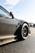 A sleek, carbon-fiber sports car with aerodynamic design is prominently featured, with a focus on its wheel and bodywork. The background shows a clear sky with an airplane flying overhead and some people standing nearby, possibly near an airport or racetrack.