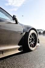 A sleek, carbon-fiber sports car with aerodynamic design is prominently featured, with a focus on its wheel and bodywork. The background shows a clear sky with an airplane flying overhead and some people standing nearby, possibly near an airport or racetrack.