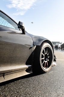 A sleek, carbon-fiber sports car with aerodynamic design is prominently featured, with a focus on its wheel and bodywork. The background shows a clear sky with an airplane flying overhead and some people standing nearby, possibly near an airport or racetrack.