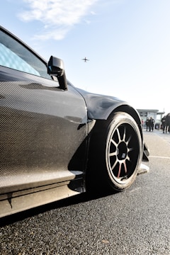 A sleek, carbon-fiber sports car with aerodynamic design is prominently featured, with a focus on its wheel and bodywork. The background shows a clear sky with an airplane flying overhead and some people standing nearby, possibly near an airport or racetrack.