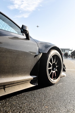 A sleek, carbon-fiber sports car with aerodynamic design is prominently featured, with a focus on its wheel and bodywork. The background shows a clear sky with an airplane flying overhead and some people standing nearby, possibly near an airport or racetrack.