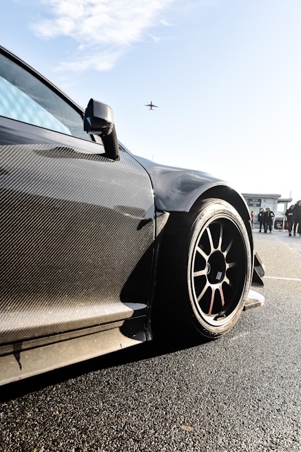 A sleek, carbon-fiber sports car with aerodynamic design is prominently featured, with a focus on its wheel and bodywork. The background shows a clear sky with an airplane flying overhead and some people standing nearby, possibly near an airport or racetrack.
