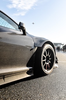 A sleek, carbon-fiber sports car with aerodynamic design is prominently featured, with a focus on its wheel and bodywork. The background shows a clear sky with an airplane flying overhead and some people standing nearby, possibly near an airport or racetrack.