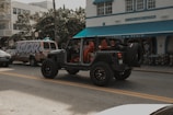 A black Jeep with open doors and red interior is parked on the street in front of a building labeled 'Shore Park Hotel'. Nearby, a van with colorful graffiti-style text reading 'AZUCAR' serves as a backdrop, and there's a café entrance with a blue awning named 'Larios on the Beach'. The scene captures a relaxed, vibrant atmosphere with a mix of vehicles and pedestrians.