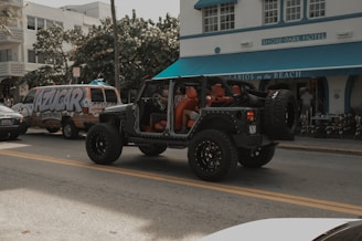 A black Jeep with open doors and red interior is parked on the street in front of a building labeled 'Shore Park Hotel'. Nearby, a van with colorful graffiti-style text reading 'AZUCAR' serves as a backdrop, and there's a café entrance with a blue awning named 'Larios on the Beach'. The scene captures a relaxed, vibrant atmosphere with a mix of vehicles and pedestrians.