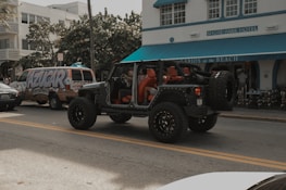 A black Jeep with open doors and red interior is parked on the street in front of a building labeled 'Shore Park Hotel'. Nearby, a van with colorful graffiti-style text reading 'AZUCAR' serves as a backdrop, and there's a café entrance with a blue awning named 'Larios on the Beach'. The scene captures a relaxed, vibrant atmosphere with a mix of vehicles and pedestrians.