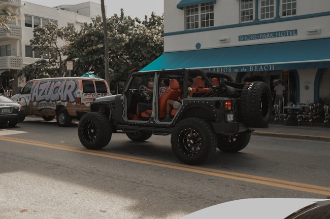 A black Jeep with open doors and red interior is parked on the street in front of a building labeled 'Shore Park Hotel'. Nearby, a van with colorful graffiti-style text reading 'AZUCAR' serves as a backdrop, and there's a café entrance with a blue awning named 'Larios on the Beach'. The scene captures a relaxed, vibrant atmosphere with a mix of vehicles and pedestrians.