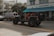 A black Jeep with open doors and red interior is parked on the street in front of a building labeled 'Shore Park Hotel'. Nearby, a van with colorful graffiti-style text reading 'AZUCAR' serves as a backdrop, and there's a café entrance with a blue awning named 'Larios on the Beach'. The scene captures a relaxed, vibrant atmosphere with a mix of vehicles and pedestrians.