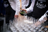 A person wearing a black apron and white shirt is pouring champagne from a bottle into one of many empty wine glasses arranged on a table. Other people wearing similar outfits are partially visible in the background.