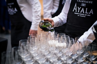 A person wearing a black apron and white shirt is pouring champagne from a bottle into one of many empty wine glasses arranged on a table. Other people wearing similar outfits are partially visible in the background.