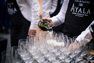 A person wearing a black apron and white shirt is pouring champagne from a bottle into one of many empty wine glasses arranged on a table. Other people wearing similar outfits are partially visible in the background.