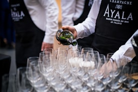 A person wearing a black apron and white shirt is pouring champagne from a bottle into one of many empty wine glasses arranged on a table. Other people wearing similar outfits are partially visible in the background.