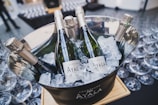 Several bottles of champagne labeled 'Ayala' are chilled in a silver ice bucket filled with ice cubes. The bucket is placed on a wooden plank and surrounded by numerous empty wine glasses.