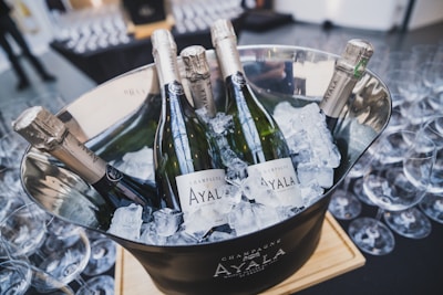 Several bottles of champagne labeled 'Ayala' are chilled in a silver ice bucket filled with ice cubes. The bucket is placed on a wooden plank and surrounded by numerous empty wine glasses.