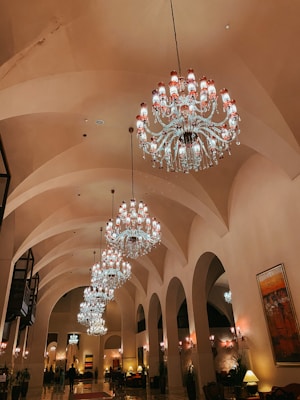 An opulent interior with multiple grand chandeliers hanging from a high, arched ceiling. The elegant lighting fixtures illuminate a spacious hall with decorative arches and ornate details. The warm lighting casts a luxurious glow over the polished floor, and a few people are seen walking and standing around.