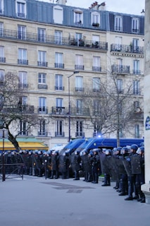 A line of police officers in full riot gear stand in formation, holding shields. Behind them are several police vehicles with flashing lights. The scene is set in an urban area with a tall residential building and bare trees.