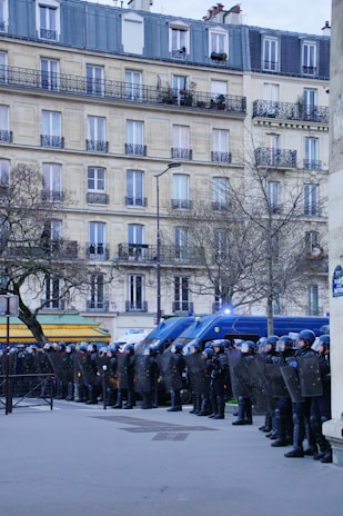 A line of police officers in full riot gear stand in formation, holding shields. Behind them are several police vehicles with flashing lights. The scene is set in an urban area with a tall residential building and bare trees.