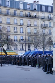 A line of police officers in full riot gear stand in formation, holding shields. Behind them are several police vehicles with flashing lights. The scene is set in an urban area with a tall residential building and bare trees.
