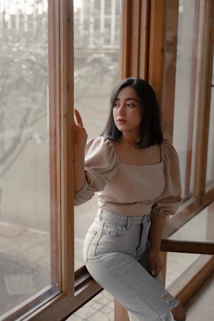 A cinematic close-up of a mature woman looking thoughtfully out a window, soft beige and nude tones surrounding her.