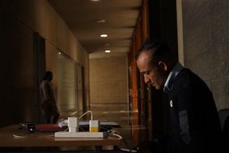 A man in a uniform is sitting at a desk in a dimly lit hallway, focused on a device in his hands. The hallway is long, with dark walls and evenly spaced lights on the ceiling. In the background, a blurred figure is walking away, adding depth to the scene. On the desk, there are a few books, a device, and some cables.