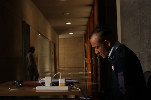 A man in a uniform is sitting at a desk in a dimly lit hallway, focused on a device in his hands. The hallway is long, with dark walls and evenly spaced lights on the ceiling. In the background, a blurred figure is walking away, adding depth to the scene. On the desk, there are a few books, a device, and some cables.