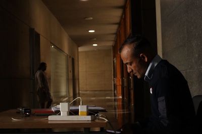 A man in a uniform is sitting at a desk in a dimly lit hallway, focused on a device in his hands. The hallway is long, with dark walls and evenly spaced lights on the ceiling. In the background, a blurred figure is walking away, adding depth to the scene. On the desk, there are a few books, a device, and some cables.