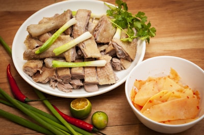 A plate of sliced meat garnished with green onions and cilantro is placed on a wooden table. Next to it is a bowl containing slices of pickled food with chili flakes. Fresh ingredients, including red and green chilies, green onions, and a small lime, are scattered around.