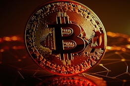 A large, close-up view of a Bitcoin coin with intricate circuit-like patterns on its surface. The background is a dark, abstract representation of a digital network with glowing orange and yellow lights.