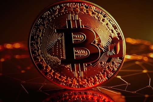 A large, close-up view of a Bitcoin coin with intricate circuit-like patterns on its surface. The background is a dark, abstract representation of a digital network with glowing orange and yellow lights.
