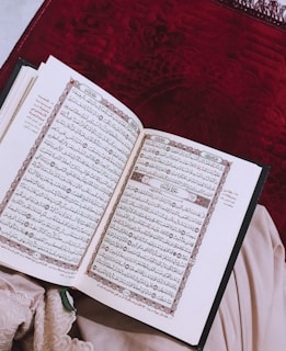 An open book displaying intricate Arabic script, likely a religious text, placed on a deep red textured fabric background. A piece of light fabric is partially visible beside the book.