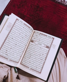 An open book displaying intricate Arabic script, likely a religious text, placed on a deep red textured fabric background. A piece of light fabric is partially visible beside the book.