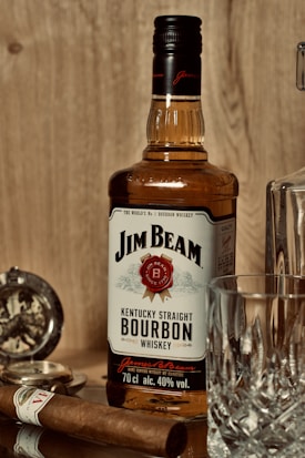 A bottle of Jim Beam Kentucky Straight Bourbon Whiskey is placed in the center. To the right, there is a crystal whiskey glass, and to the left, a cigar and a pocket watch are also visible. The setting has a warm and vintage atmosphere with a wooden background.