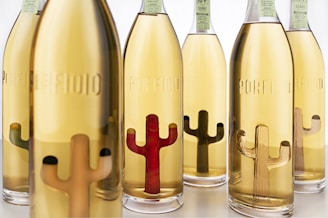 A collection of glass bottles filled with a golden liquid is arranged closely together. Each bottle features an embossed motif of a cactus shape in varying colors such as brown and red, which adds a unique design element. The bottles are labeled with 'Porfidio' and have a green label on top.