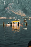 Wooden boats with colorful canopies are floating on a calm body of water. In the background, there are large mountains with a village and shoreline visible near the base. The reflection of the boats and surrounding scenery is visible on the water.