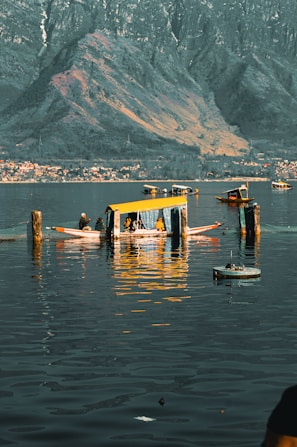 Wooden boats with colorful canopies are floating on a calm body of water. In the background, there are large mountains with a village and shoreline visible near the base. The reflection of the boats and surrounding scenery is visible on the water.