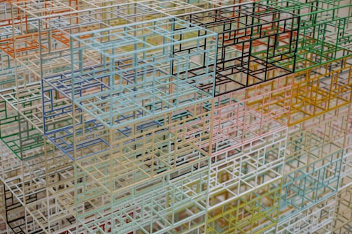 A complex and intricate structure composed of numerous intersecting metal or wireframe cubes in varying colors. The arrangement appears in a geometric pattern with a sense of spatial depth and layering.
