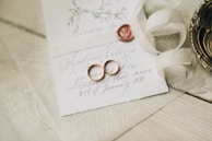 A close-up of an elegantly arranged wedding invitation featuring handwritten calligraphy. Two gold wedding rings are placed on the card alongside a red wax seal. A cream-colored ribbon is artistically draped nearby, and a decorative metal dish is partially visible.