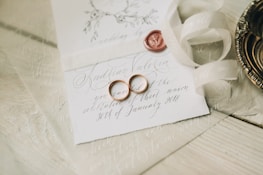 A close-up of an elegantly arranged wedding invitation featuring handwritten calligraphy. Two gold wedding rings are placed on the card alongside a red wax seal. A cream-colored ribbon is artistically draped nearby, and a decorative metal dish is partially visible.