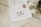 A close-up of an elegantly arranged wedding invitation featuring handwritten calligraphy. Two gold wedding rings are placed on the card alongside a red wax seal. A cream-colored ribbon is artistically draped nearby, and a decorative metal dish is partially visible.