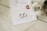 A close-up of an elegantly arranged wedding invitation featuring handwritten calligraphy. Two gold wedding rings are placed on the card alongside a red wax seal. A cream-colored ribbon is artistically draped nearby, and a decorative metal dish is partially visible.